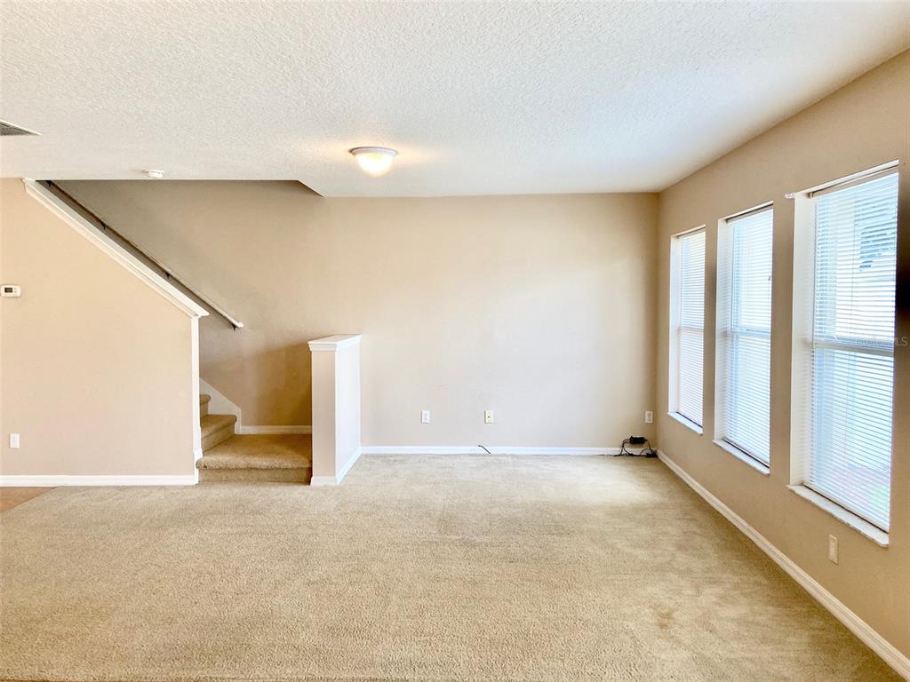 An empty living room features beige carpet, tan walls, a staircase, and three large windows — 2539 AVENTURINE STREET, KISSIMMEE, FL