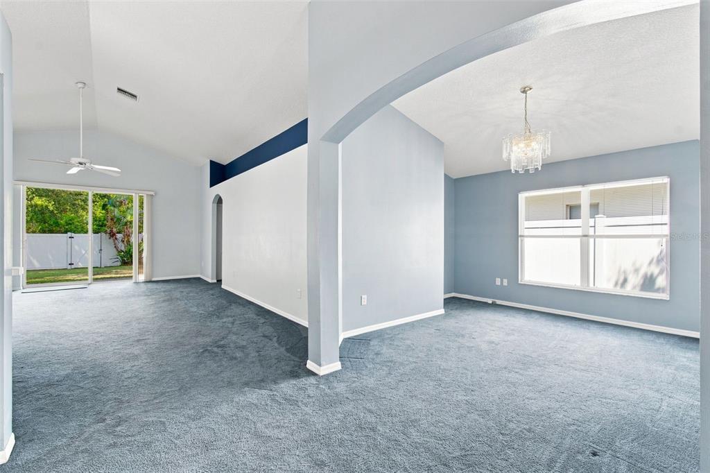 An open-concept living and dining area features blue carpeting, vaulted ceilings, and a crystal chandelier — 3838 BELLEWATER BLVD, RIVERVIEW, FL