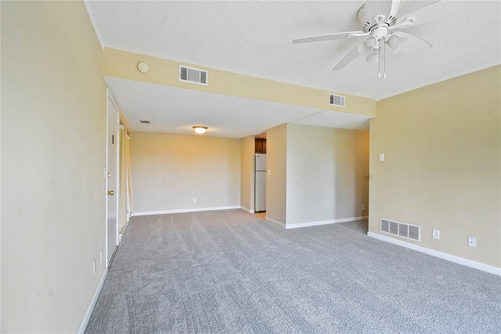 An empty living room features gray carpeting, a ceiling fan, and an adjacent kitchen area — 40 CLUB HOUSE DRIVE UNIT 207, PALM COAST, FL
