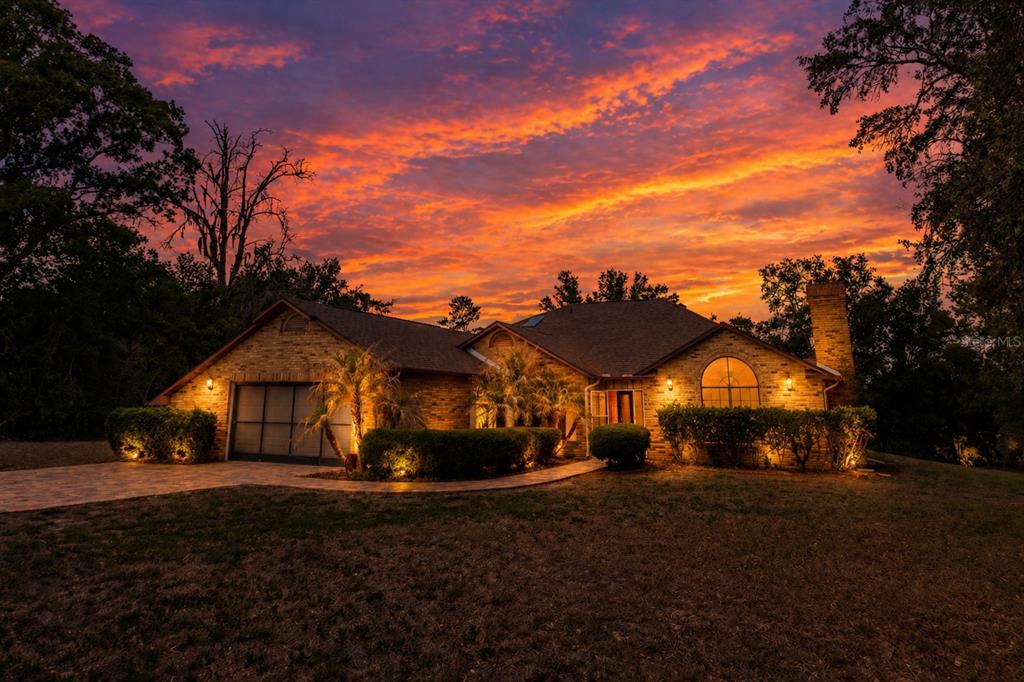 A brick house with a garage and arched window stands under a vibrant sunset sky — 14692 LINDEN DRIVE, SPRING HILL, FL