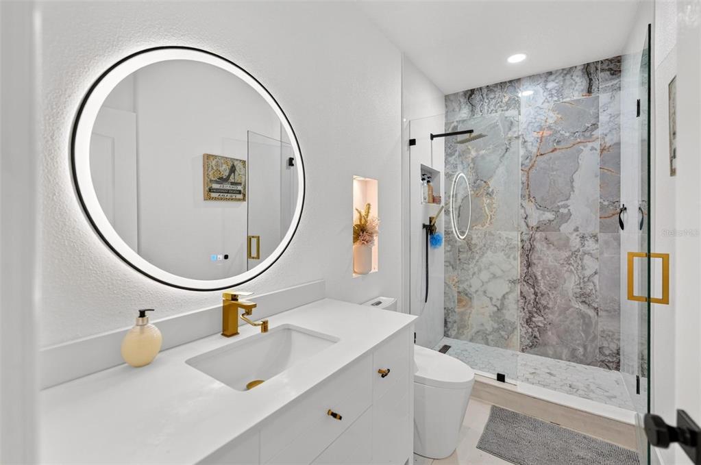 A modern bathroom features a round LED mirror, white vanity, gold faucet, and a marble-tiled shower — 3016 CAMINO REAL DRIVE S, KISSIMMEE, FL