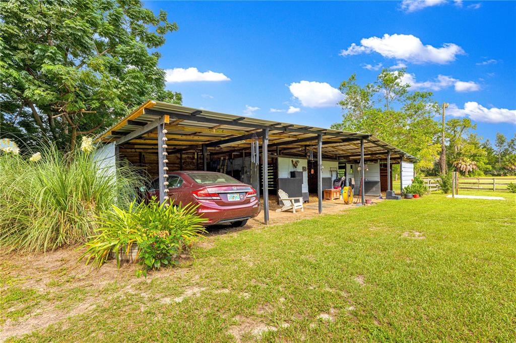 A red sedan is parked under a metal-roofed barn structure on a grassy lot — 8221 SE 126TH PLACE, BELLEVIEW, FL