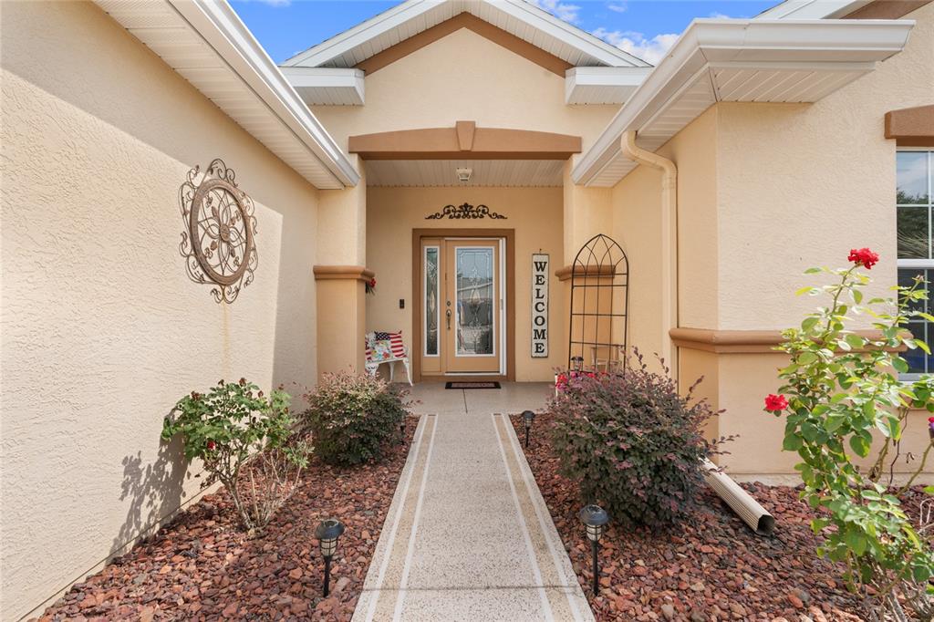 A tan stucco house exterior features a concrete walkway leading to a glass-paneled front door — 9215 SE 118TH LOOP, SUMMERFIELD, FL