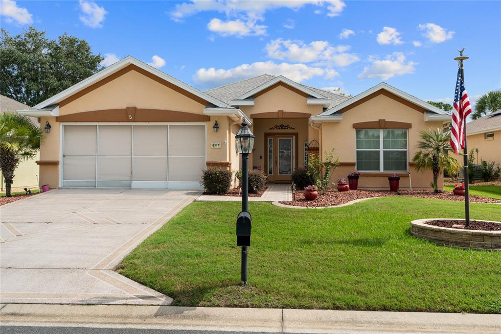 A single-story tan house with a two-car garage, a front lawn, and an American flag — 9215 SE 118TH LOOP, SUMMERFIELD, FL