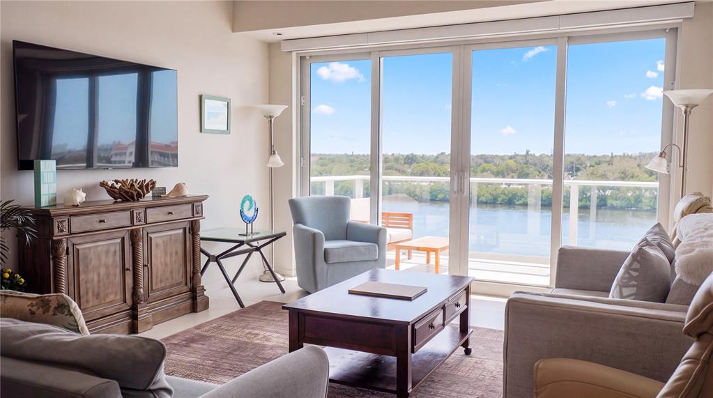A living room features a wooden console, coffee table, armchairs, and a balcony overlooking water — 1020 SUNSET POINT ROAD UNIT 512, CLEARWATER, FL