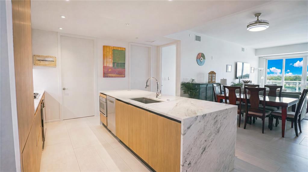 A modern kitchen features a marble-topped island, light wood cabinetry, and an adjacent dining area — 1020 SUNSET POINT ROAD UNIT 512, CLEARWATER, FL