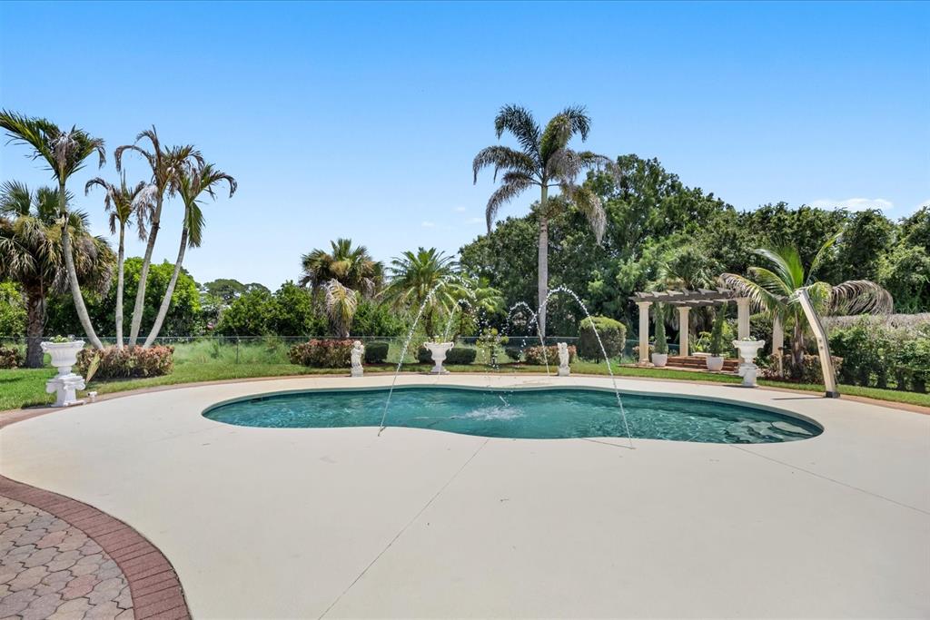 A kidney-shaped swimming pool with fountains sits in a backyard surrounded by palm trees — 1819 SE ELROSE STREET, PORT ST LUCIE, FL