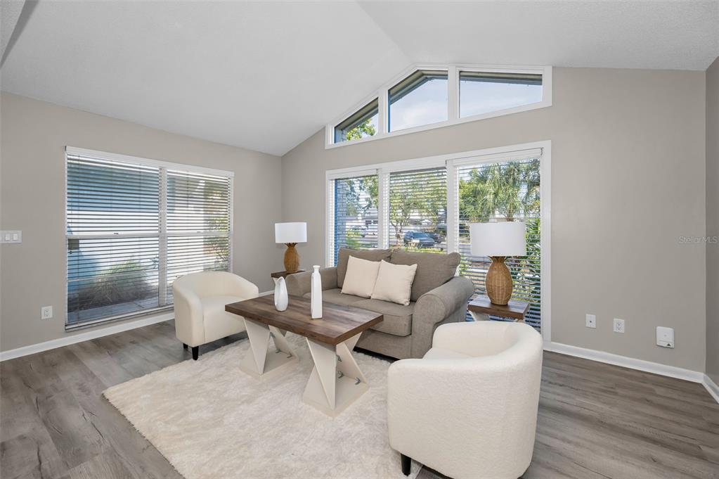 A living room with vaulted ceilings, large windows, gray wood-look flooring, and neutral-toned furniture — 7404 CLEARVIEW DRIVE, TAMPA, FL