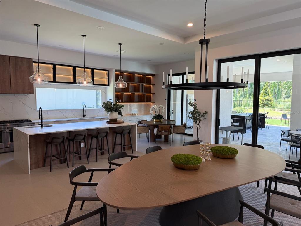 A modern open-concept kitchen and dining area featuring a large island, wooden cabinetry, and patio access — 15544 PENDIO DRIVE, BELLA COLLINA, FL