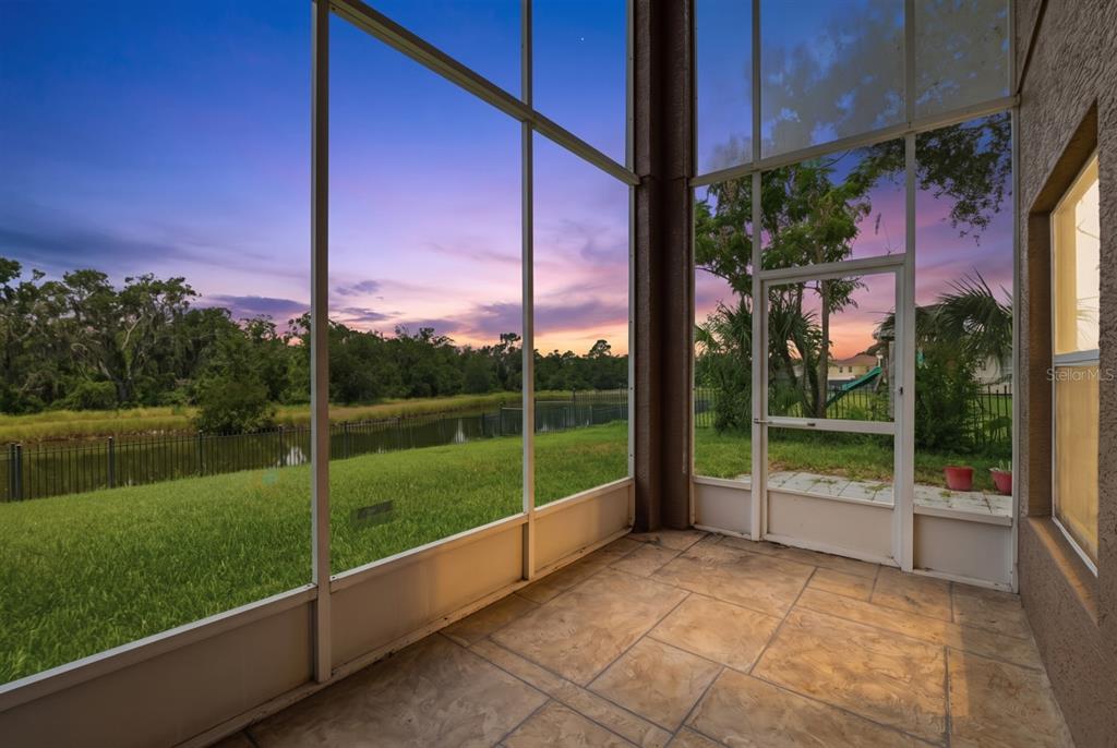 A screened-in patio with tile flooring overlooks a grassy yard, a pond, and sunset skies — 14722 HERONGLEN DRIVE, LITHIA, FL
