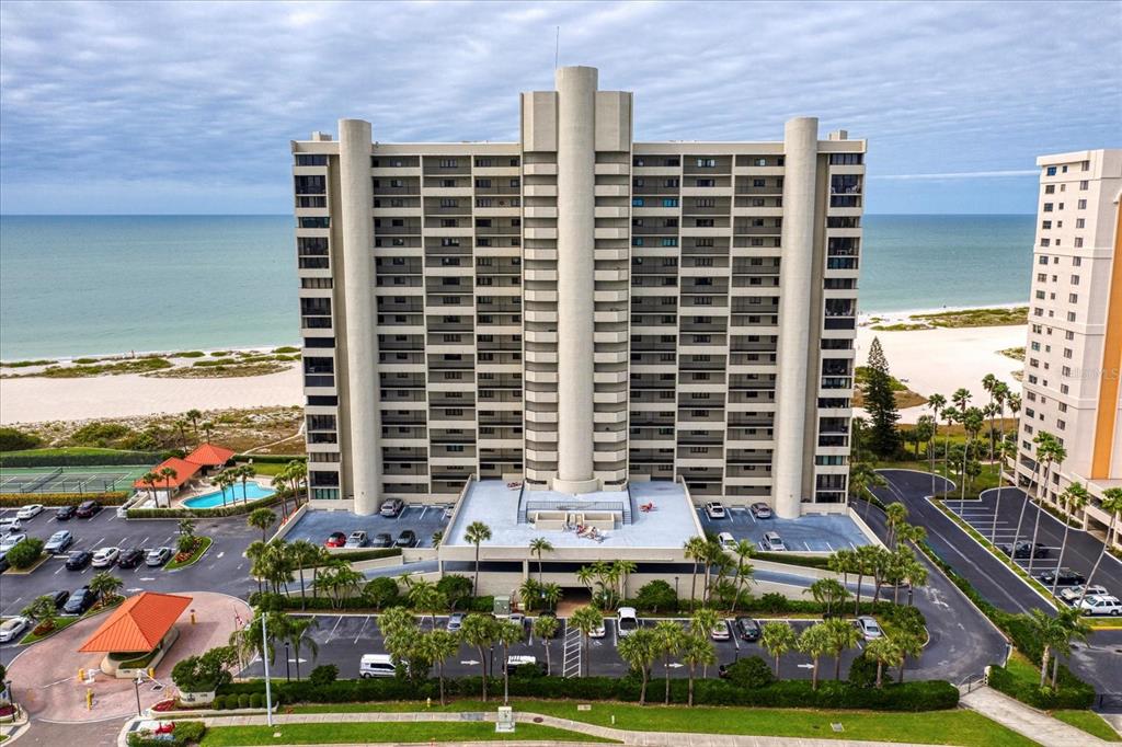 An aerial view shows a tall beachfront condominium building with a parking lot and pool — 1290 GULF BOULEVARD UNIT 1406, CLEARWATER, FL