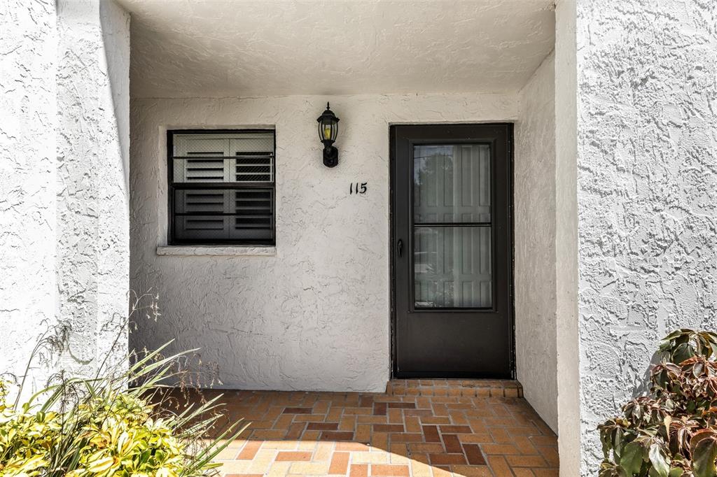 A white stucco building entrance features a brick floor, a black door, and a window — 1730 MANASOTA BEACH ROAD UNIT 115, ENGLEWOOD, FL