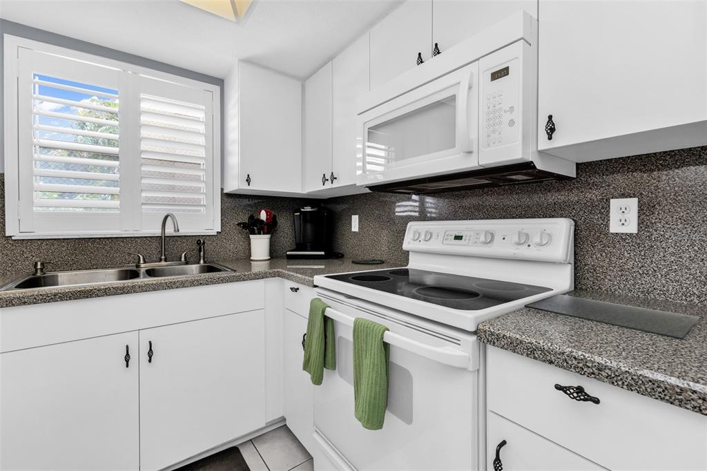 A white kitchen features granite countertops, white cabinetry, a window with shutters, and white appliances — 1730 MANASOTA BEACH ROAD UNIT 115, ENGLEWOOD, FL
