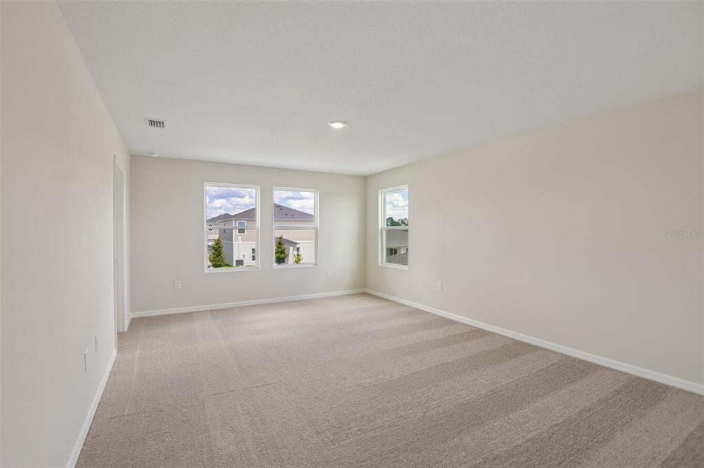 An empty bedroom features beige walls, neutral carpeting, and three windows overlooking a suburban neighborhood — 35033 ARBOR RETREAT DRIVE, DADE CITY, FL
