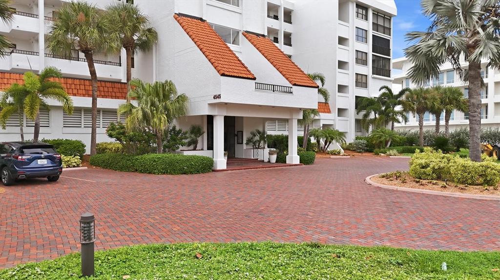 A multi-story white building with orange tiled roof accents features a brick-paved circular driveway — 4545 GULF OF MEXICO DRIVE UNIT 311, LONGBOAT KEY, FL