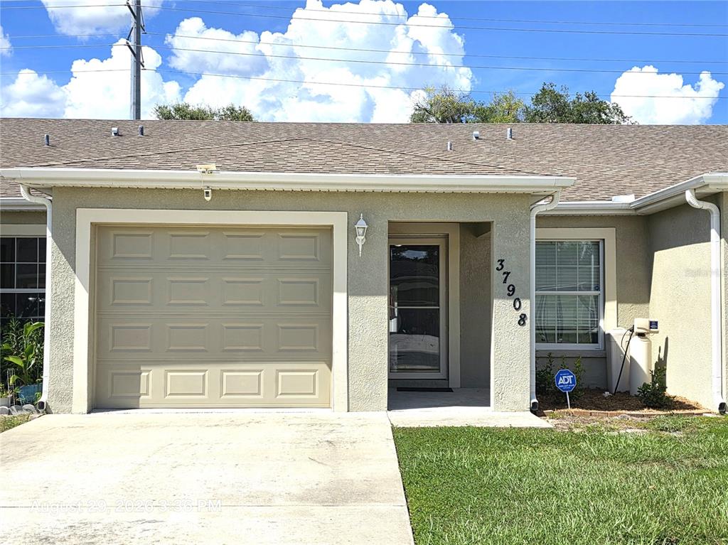 The exterior of a single-story house features a tan garage, a front entrance, and a window — 37908 PRAIRIE ROSE LOOP, ZEPHYRHILLS, FL