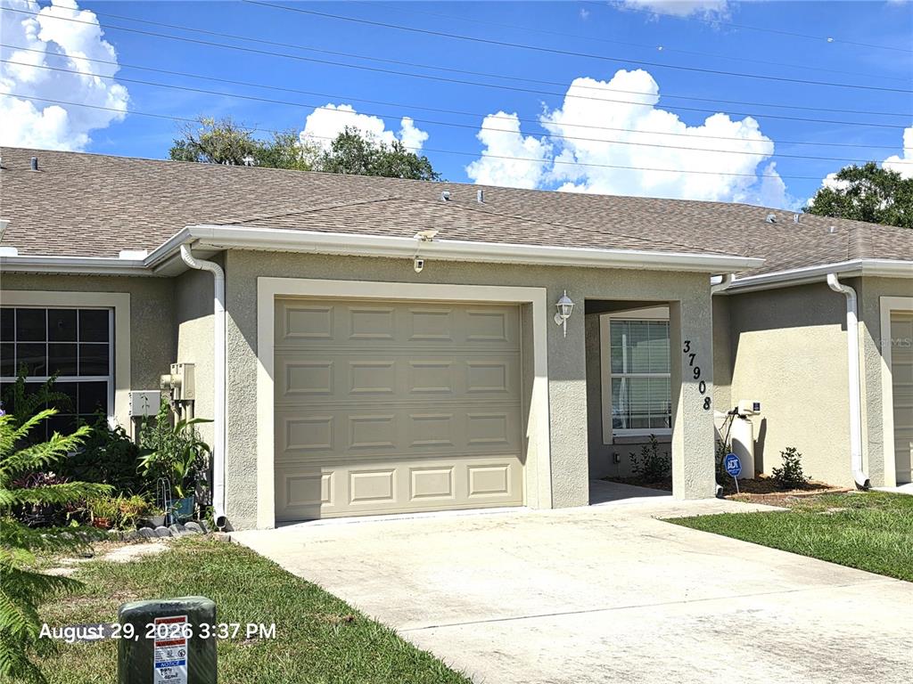 A single-story residential building features a tan stucco exterior, a one-car garage, and a concrete driveway — 37908 PRAIRIE ROSE LOOP, ZEPHYRHILLS, FL