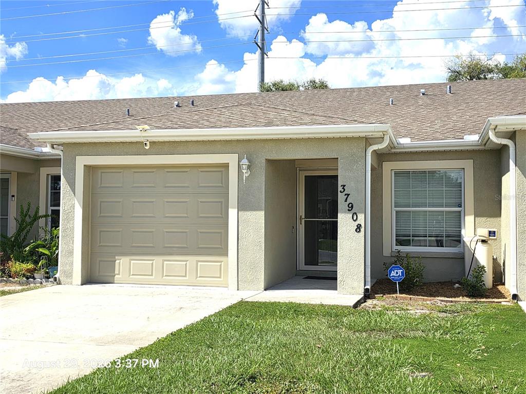 A single-story house exterior features a tan stucco facade, a garage, and a front entrance — 37908 PRAIRIE ROSE LOOP, ZEPHYRHILLS, FL