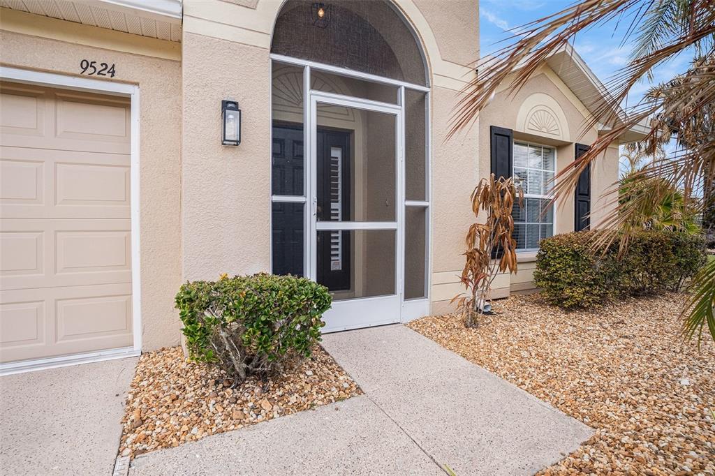 A beige house exterior features a garage, a screened front entryway, and a gravel yard — 9524 ROLLING CIRCLE, SAN ANTONIO, FL