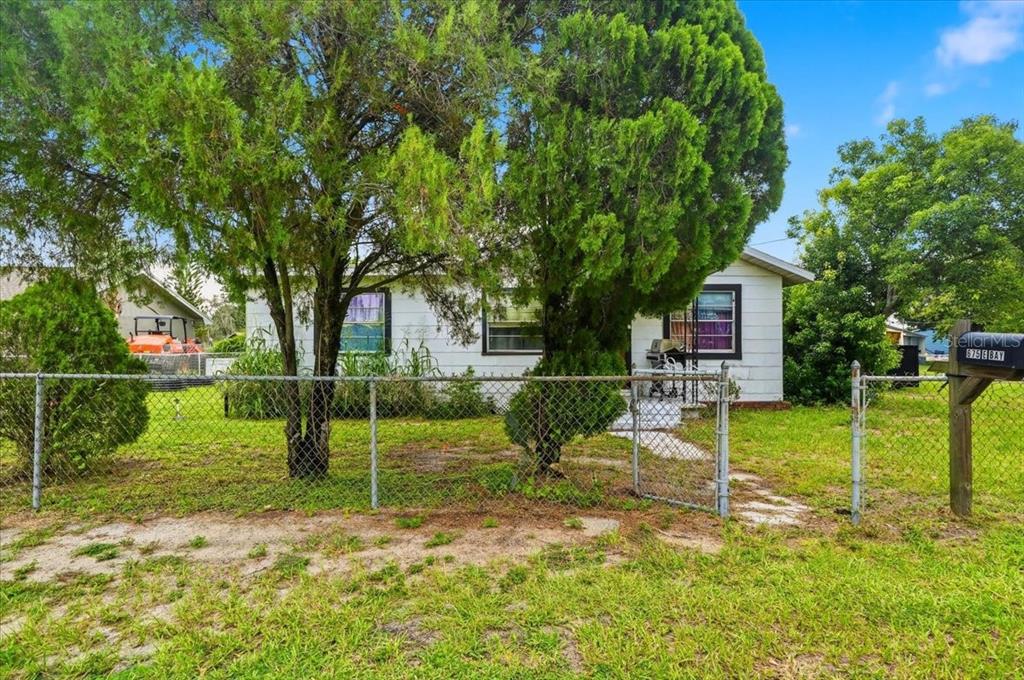 A white single-story house is viewed from the street behind a chain-link fence and trees — 675 E BAY AVENUE, EAGLE LAKE, FL
