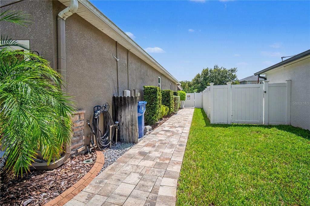 A paved walkway runs alongside a house exterior with a white fence and green lawn — 18756 BURNDALL COURT, LAND O LAKES, FL
