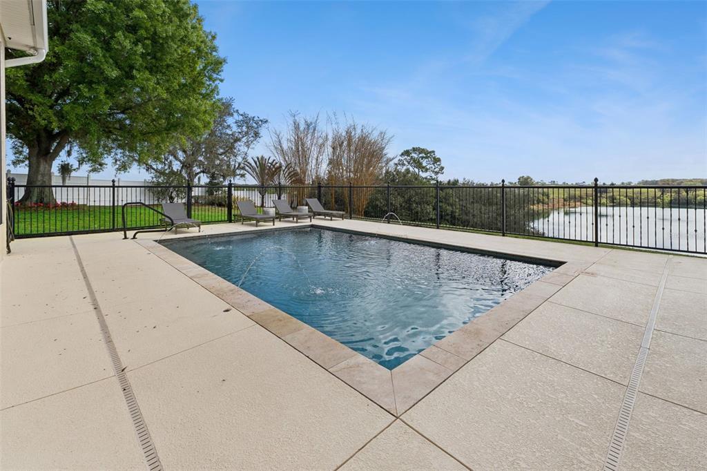 A rectangular swimming pool with fountains sits on a patio overlooking a lake and trees — 3231 HARBOR BEACH DRIVE, LAKE WALES, FL