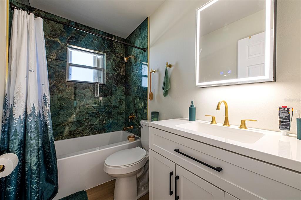 A bathroom features a green marble-tiled shower, a white vanity, and a backlit mirror — 6401 CEDARBROOK DRIVE S, PINELLAS PARK, FL
