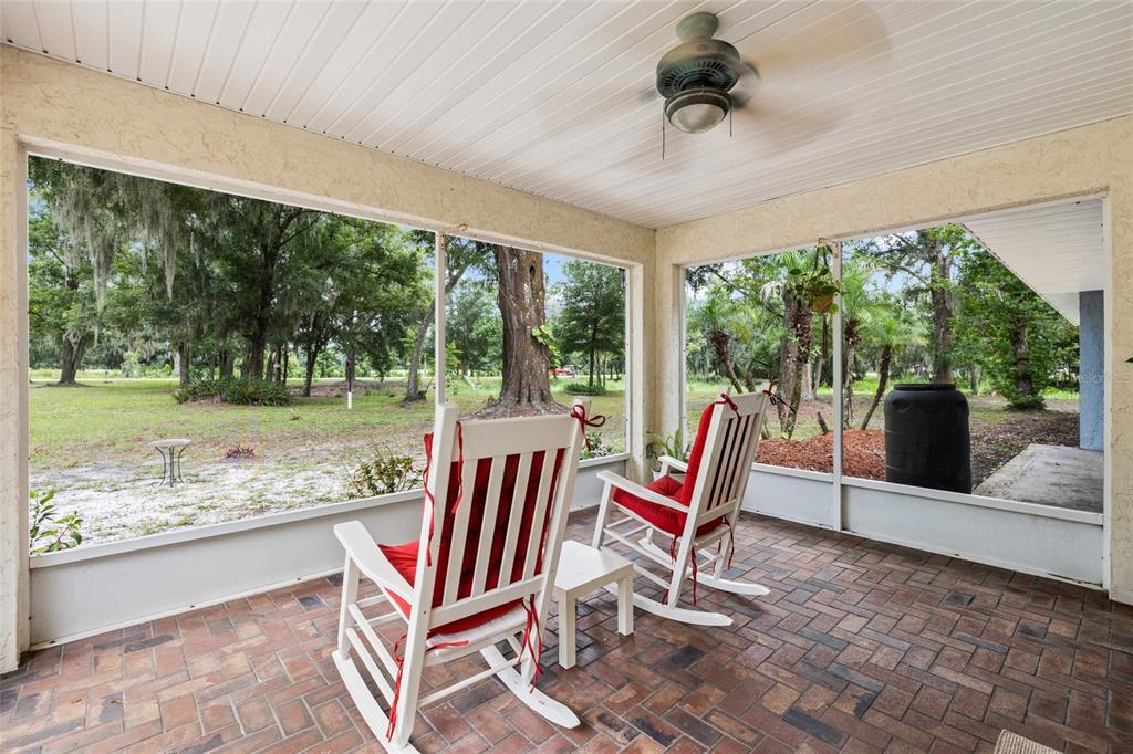 A screened-in porch with brick flooring, two rocking chairs, and a ceiling fan overlooking trees — 7402 W KNIGHTS GRIFFIN ROAD, PLANT CITY, FL