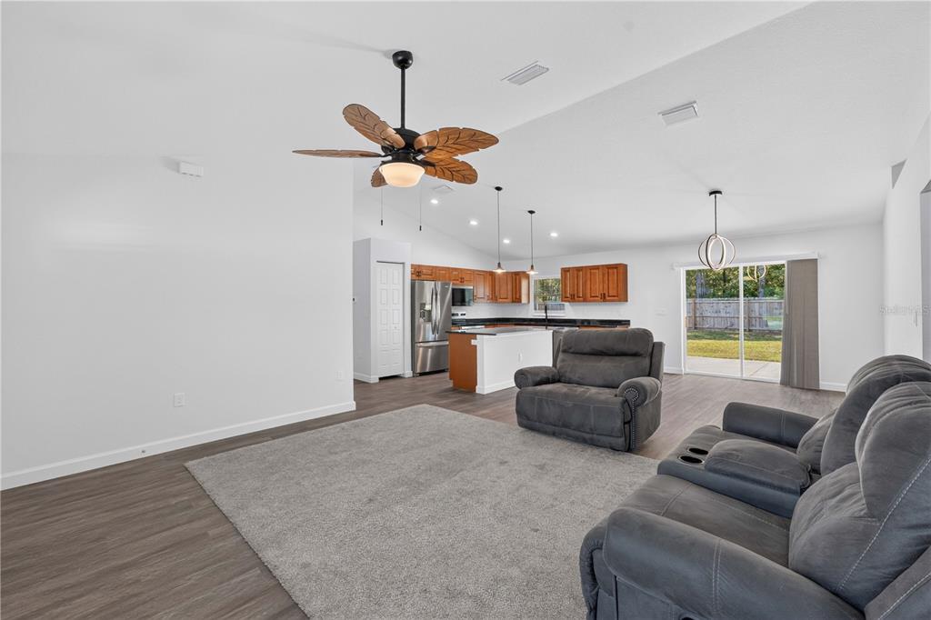 An open-concept living room with a gray rug, gray reclining furniture, and a kitchen beyond — 138 FISHER RUN, OCKLAWAHA, FL