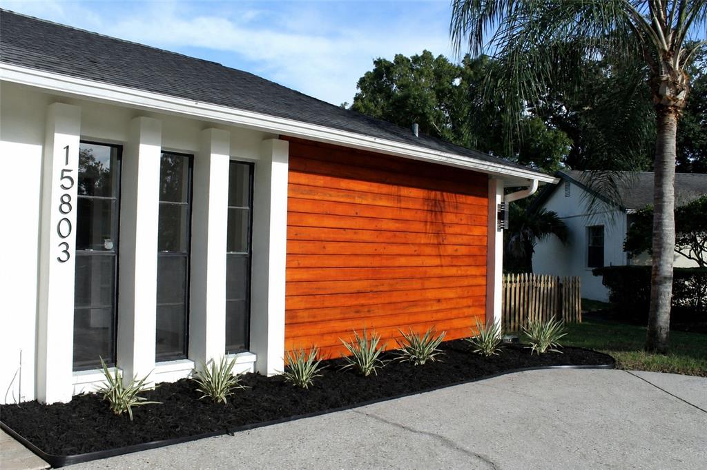 A house exterior features white walls, a wood-paneled section, black-framed windows, and a landscaped garden — 15803 IRONWARE PLACE, TAMPA, FL