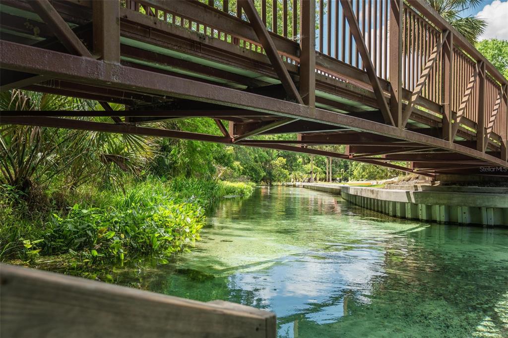 A metal pedestrian bridge spans a clear, shallow canal lined with lush vegetation and concrete walls — KELLY PARK ROAD E, APOPKA, FL