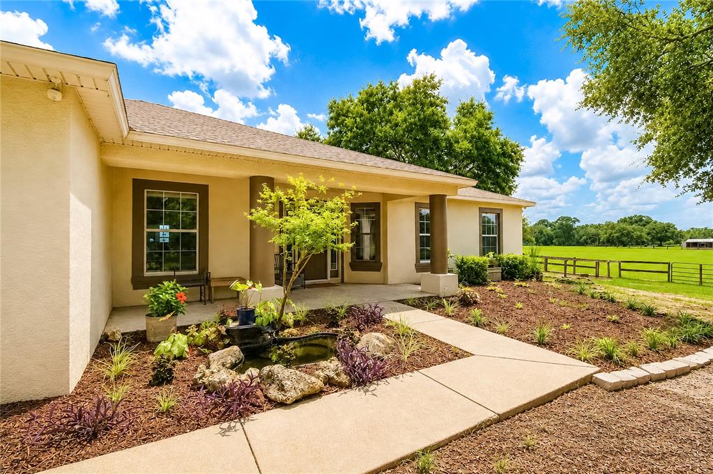 A single-story house exterior features a covered porch, a small pond, and a grassy field — 10525 SW 129TH TERRACE ROAD, DUNNELLON, FL