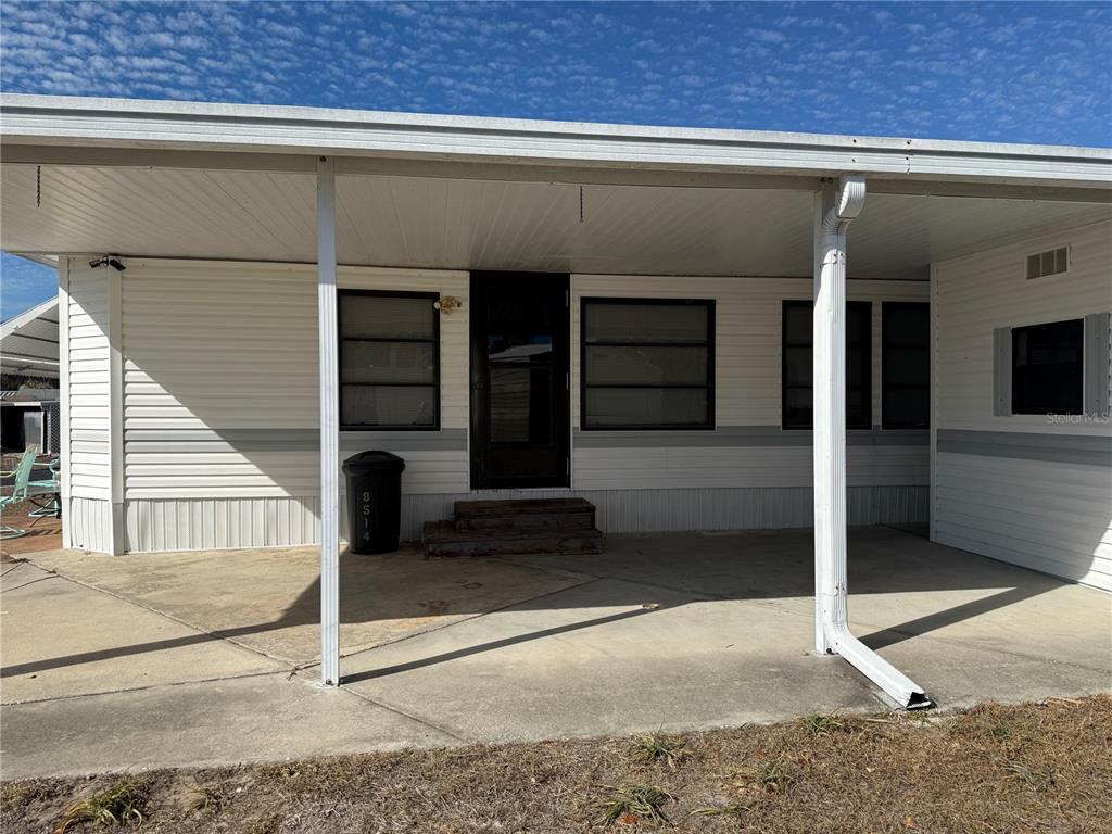 A white mobile home exterior features a covered concrete patio and a dark front door — 9514 BLACK BEAR DRIVE, WEEKI WACHEE, FL