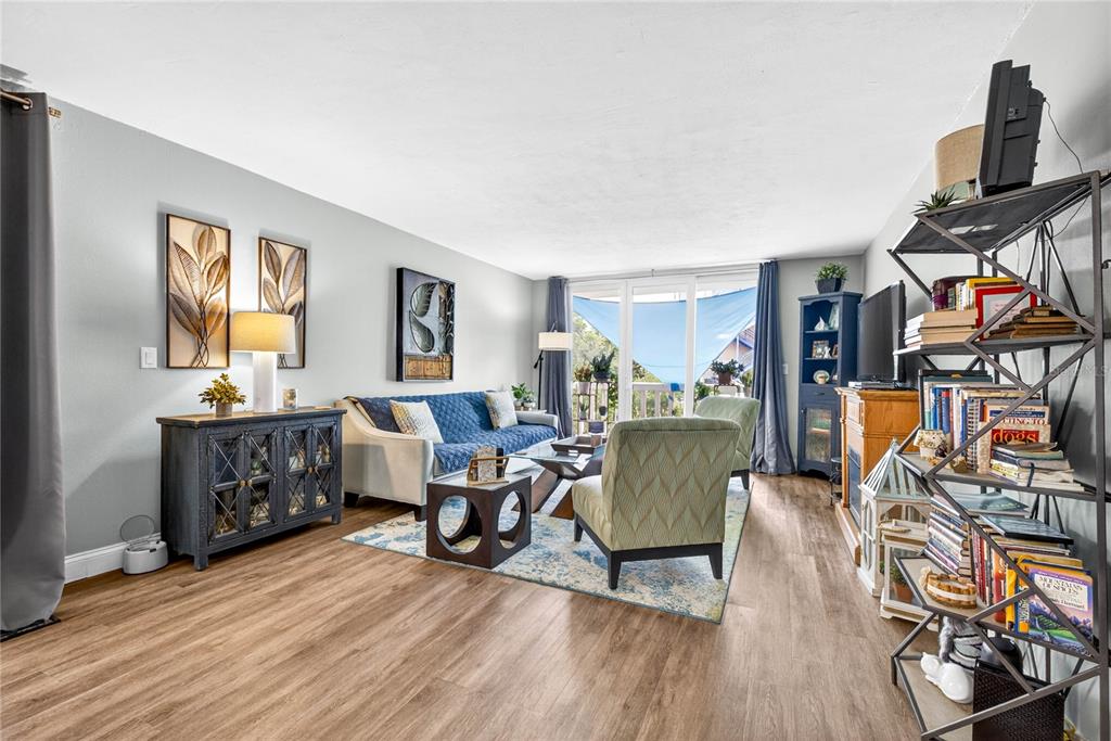 A living room features wood-look flooring, a blue sofa, patterned armchairs, and a metal bookshelf — 2623 SEVILLE BOULEVARD UNIT 207, CLEARWATER, FL