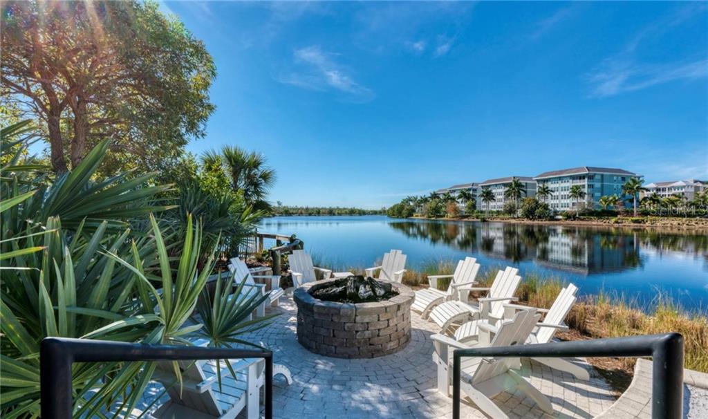 A stone fire pit sits on a paved patio overlooking a lake with apartment buildings nearby — 395 ARUBA CIRCLE UNIT 203, BRADENTON, FL