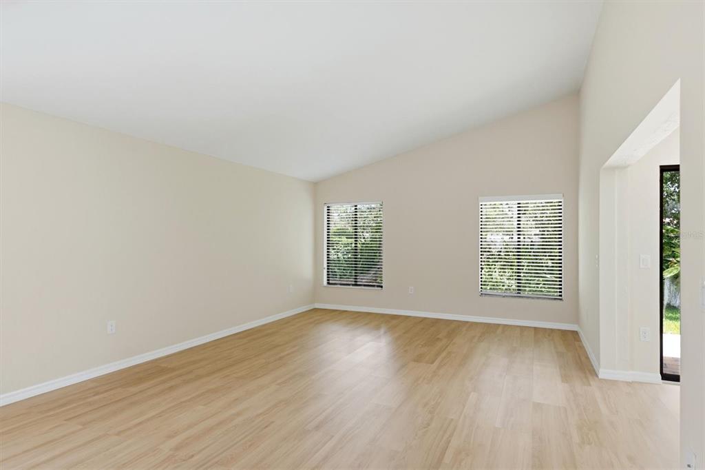 An empty living room features light-colored walls, wood-look flooring, and two windows with blinds — 6403 PELICAN DRIVE, BRADENTON, FL