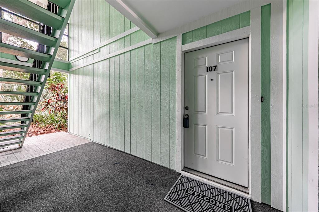 A white front door labeled 107 sits next to a green wall and outdoor staircase — 3295 FOX CHASE CIRCLE N UNIT 107, PALM HARBOR, FL