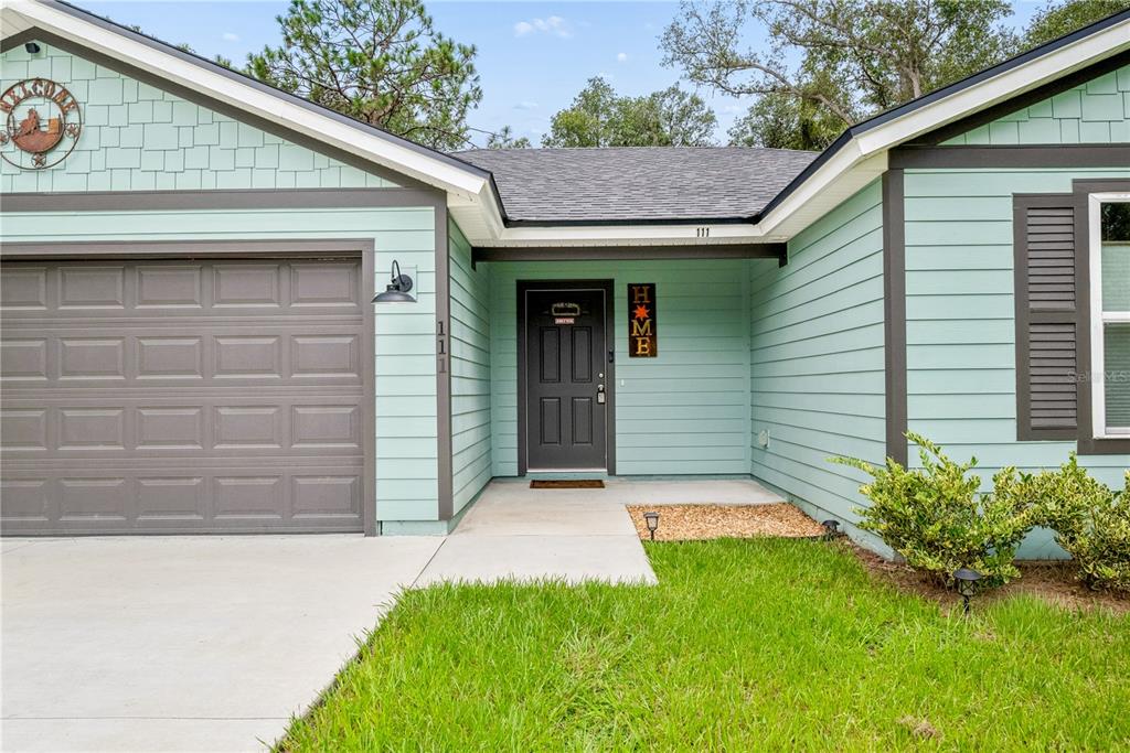 A light green house exterior features a dark gray garage door and front door — 111 BREEZEWAY AVENUE, SATSUMA, FL