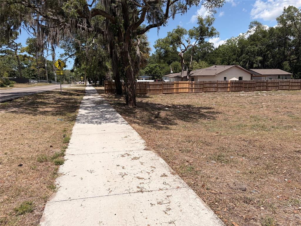 A concrete sidewalk runs alongside a vacant, grassy lot with a wooden fence and trees — HICKS ROAD, HUDSON, FL