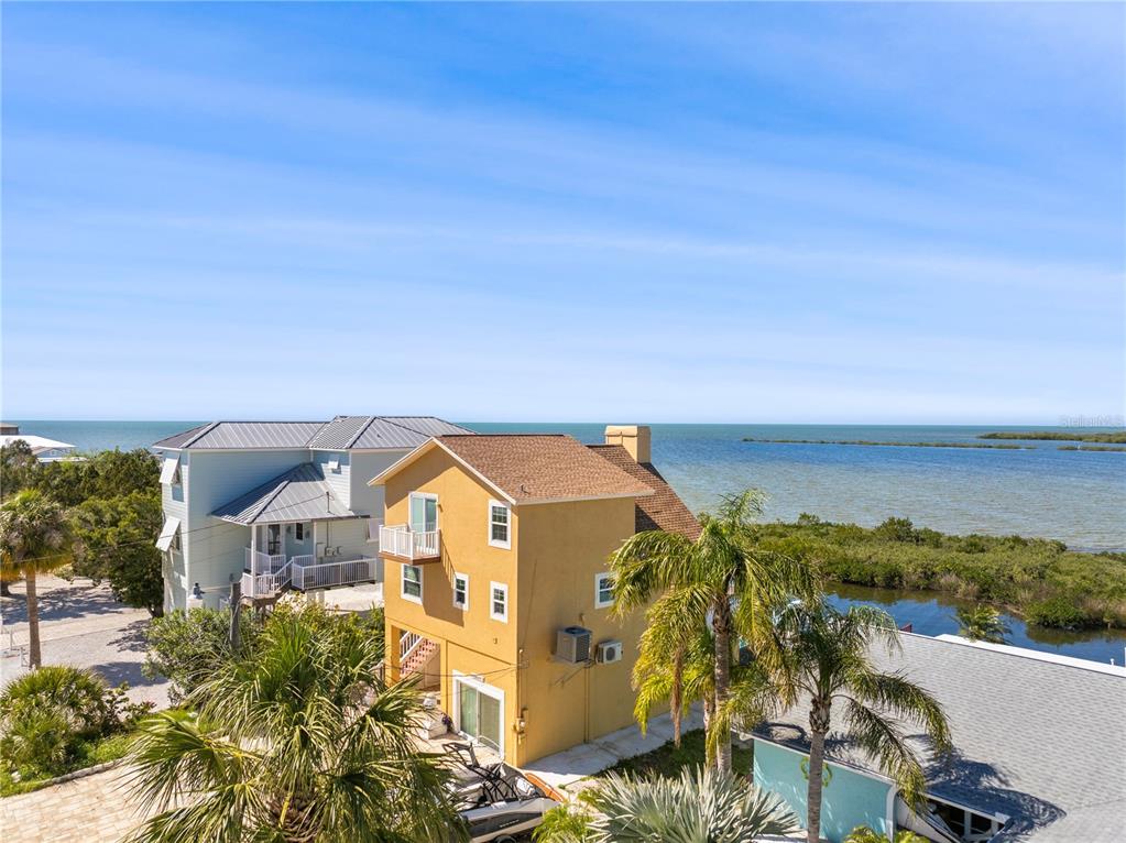 An elevated yellow house stands near a coastal waterway with palm trees and blue skies — 6633 DRIFTWOOD DRIVE, HUDSON, FL