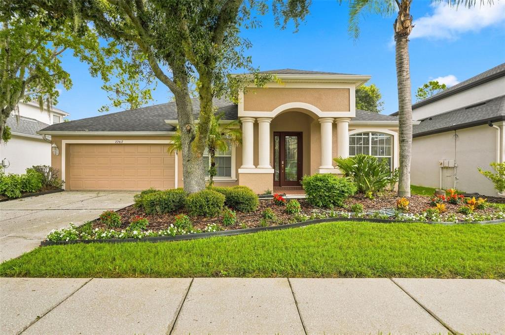 A single-story house with a tan stucco exterior, a two-car garage, and a columned entryway — 27417 SILVER THATCH DRIVE, WESLEY CHAPEL, FL