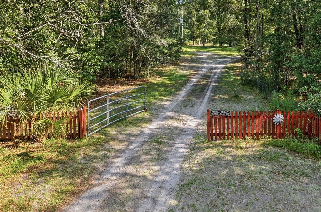 A dirt driveway leads through an open metal gate flanked by red wooden picket fencing — 24628 NW 84TH PLACE, ALACHUA, FL