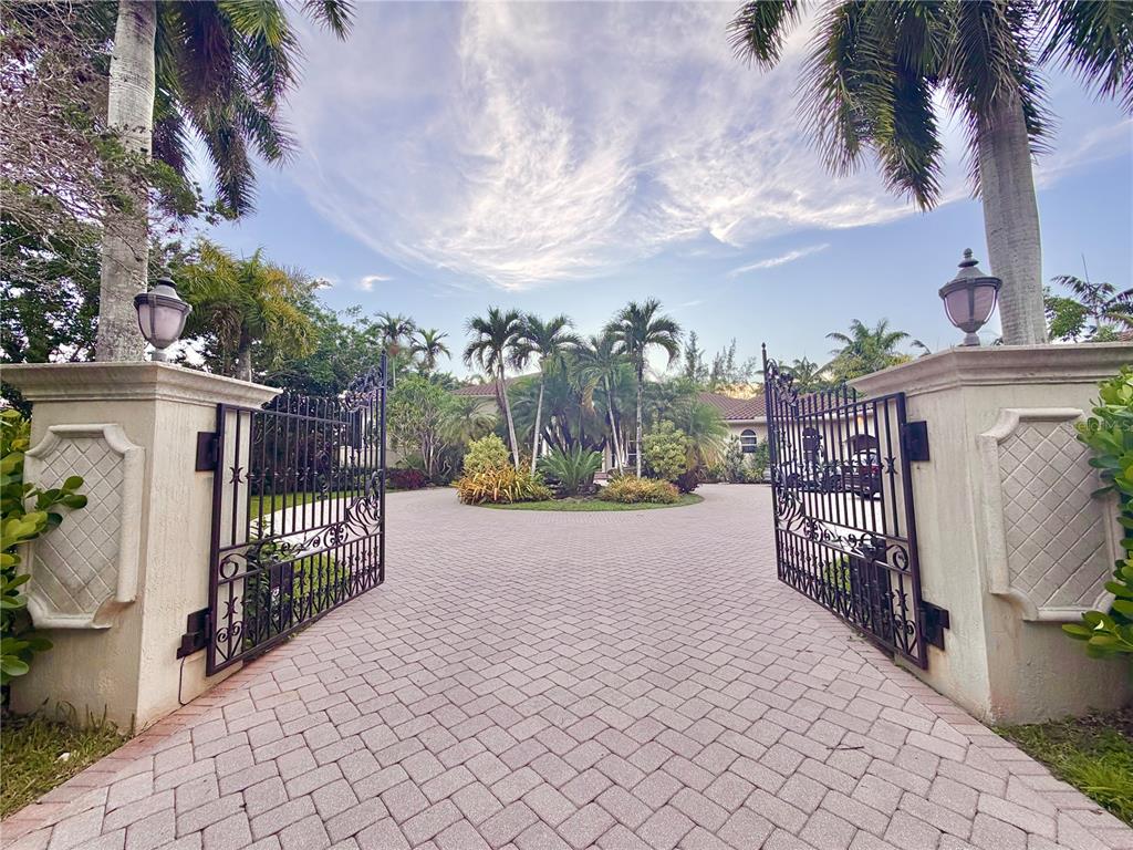 A paved driveway leads through an open wrought iron gate toward a landscaped tropical garden — 471 RANCH ROAD, WESTON, FL