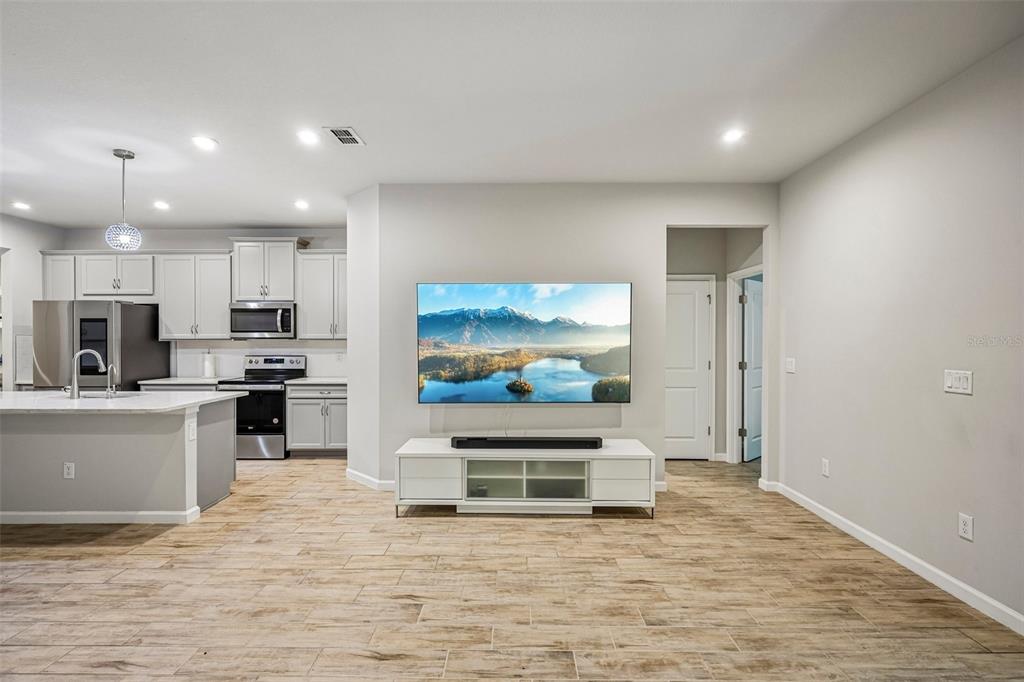 A living room with light wood-look tile flooring, a wall-mounted television, and an adjacent kitchen — 19405 MONARCH WIND WAY, LUTZ, FL
