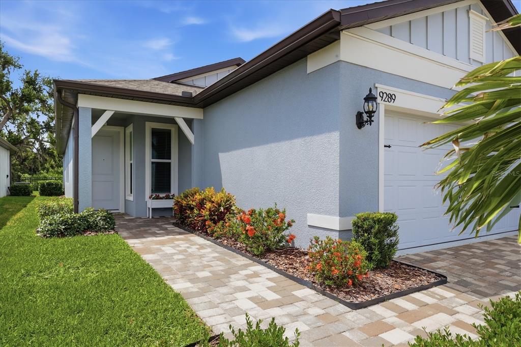 A light blue house exterior features a paved walkway, a garage, and landscaped garden beds — 9289 BALLASTER POINTE LOOP, PARRISH, FL