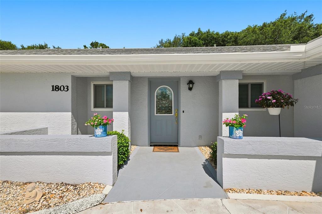 The exterior of a single-story house features a gray front door and a paved walkway — 1803 NEW BEDFORD DRIVE, SUN CITY CENTER, FL