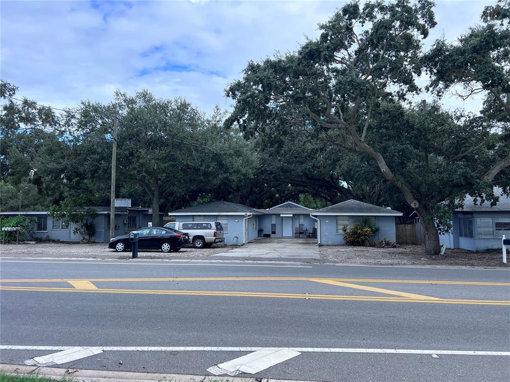 A single-story, light blue residential building sits behind a road with large oak trees — 1302 SUNSET PT RD, CLEARWATER, FL