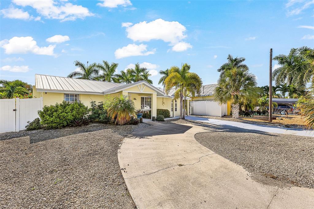 A yellow single-story house with a metal roof, gravel yard, and a concrete driveway — 12262 BOAT SHELL DR, MATLACHA ISLES, FL