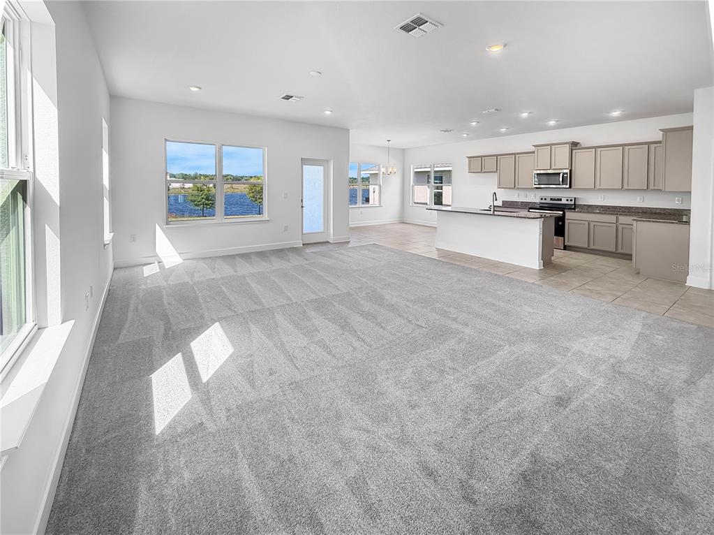 An open-concept living room with gray carpet flows into a kitchen with light-colored cabinetry — 7575 BENT TREE LOOP, HARMONY, FL