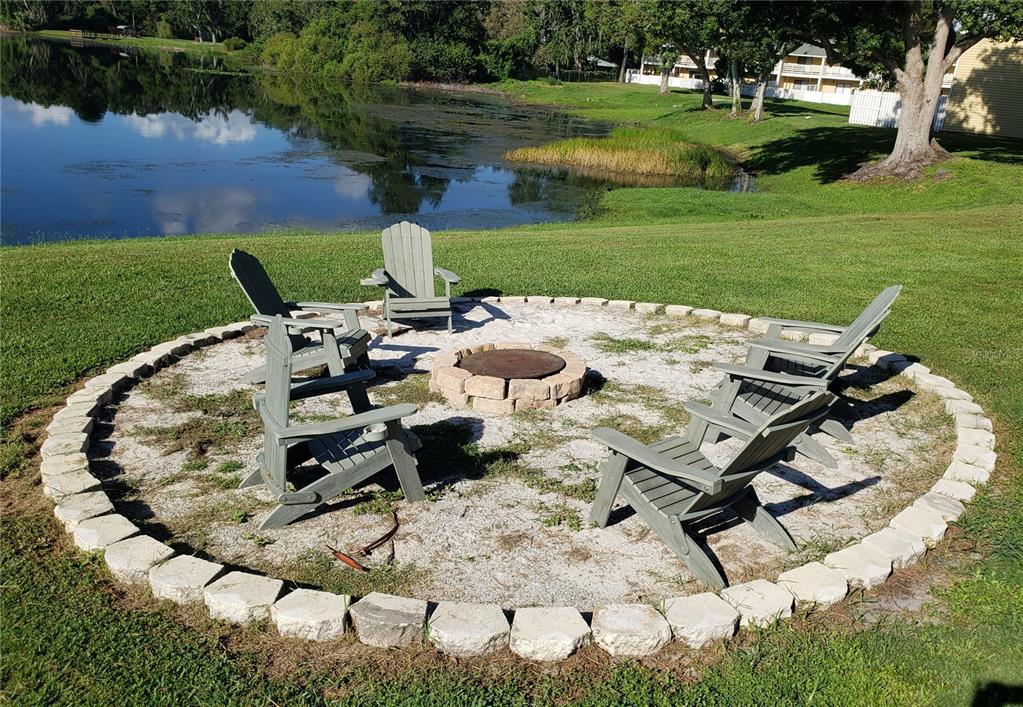 A circular stone fire pit area with Adirondack chairs sits on a lawn by a lake — 22706 GAGE LOOP UNIT 33, LAND O LAKES, FL